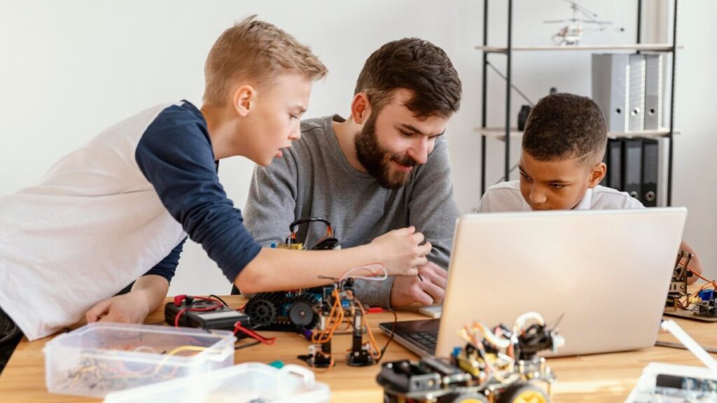 Adult and two boys engaging with electronic gadgets and a laptop at a wooden table.